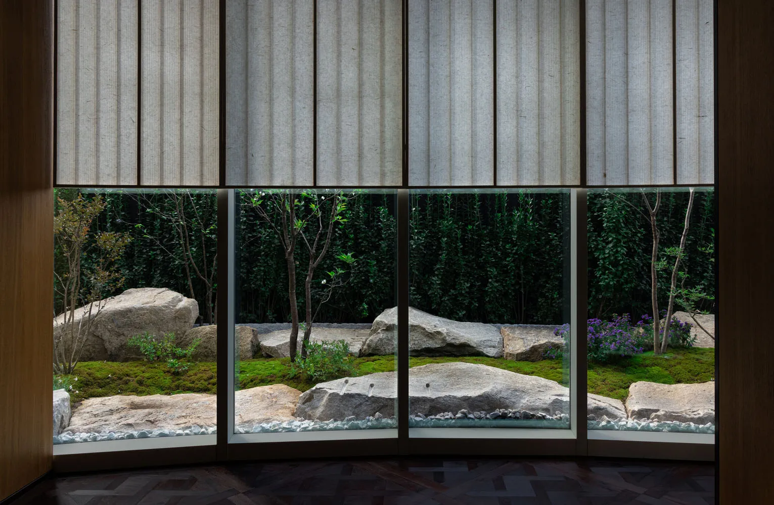 A tranquil indoor space with a large window offering a view of a garden with a pond, rocks, and plants, surrounded by trees, and natural light pouring in