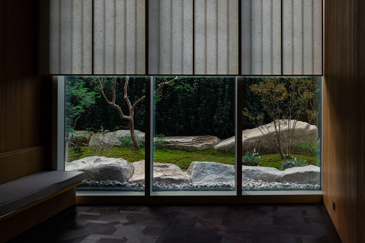 A cozy indoor space with a large window, rock wall, potted plants, and vases, offering a view of a garden outside