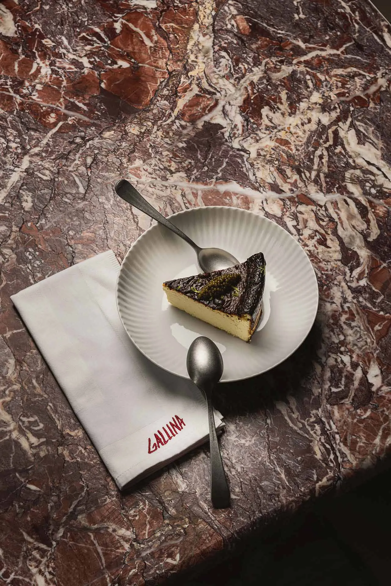 A marble tabletop with a slice of cake on a white plate, two spoons, and an elegant indoor dining setting