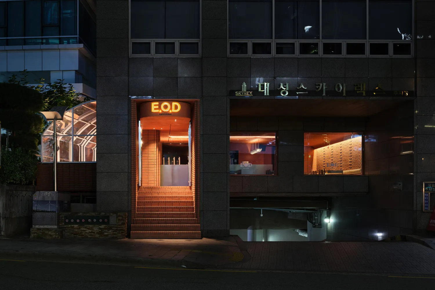 A modern Asian restaurant at night with a brick exterior, glass windows, and a warm interior atmosphere, featuring dining tables, chairs, and bottles