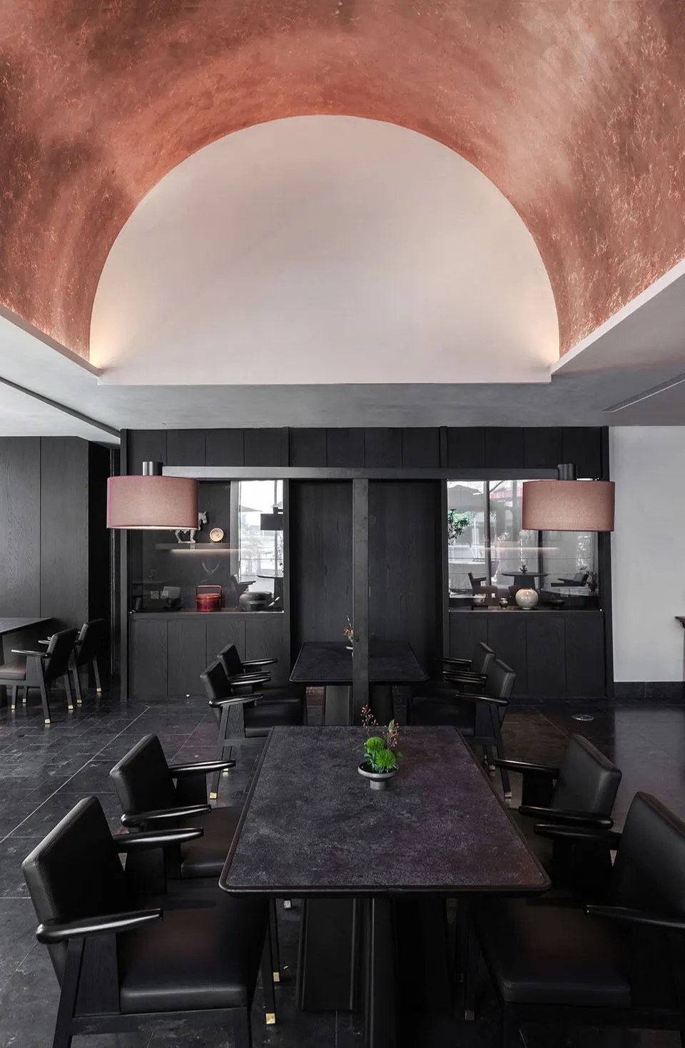 A modern restaurant dining area with a large black table surrounded by multiple chairs and a potted plant, creating a comfortable and inviting atmosphere