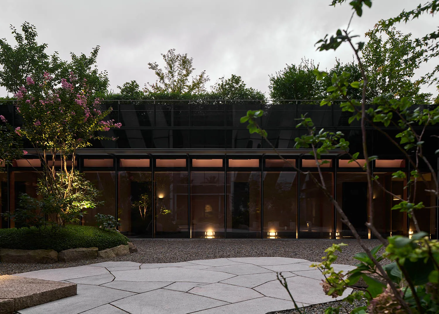 A modern building with a glass facade surrounded by trees and bushes, featuring large windows that allow natural light into the interior spaces