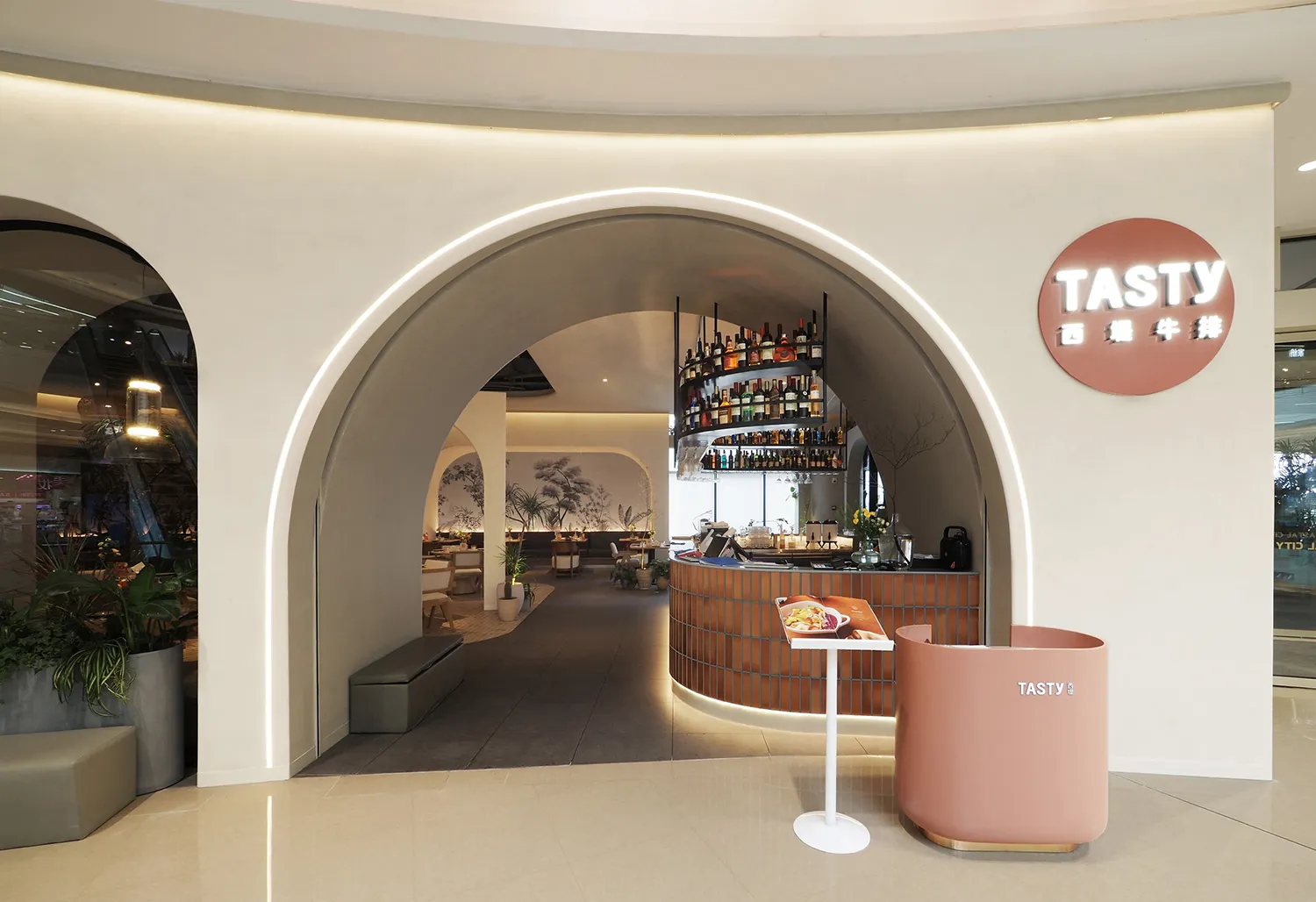 Modern restaurant interior with a bar area, potted plants, and a collection of bottles on shelves behind the counter