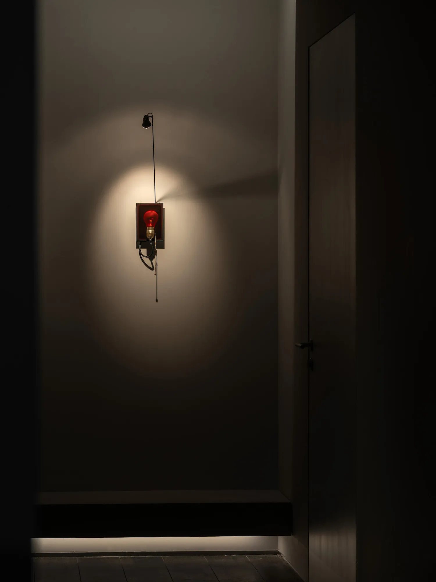 A dimly lit hallway with a door and a fire alarm with a red light, mounted to the wall