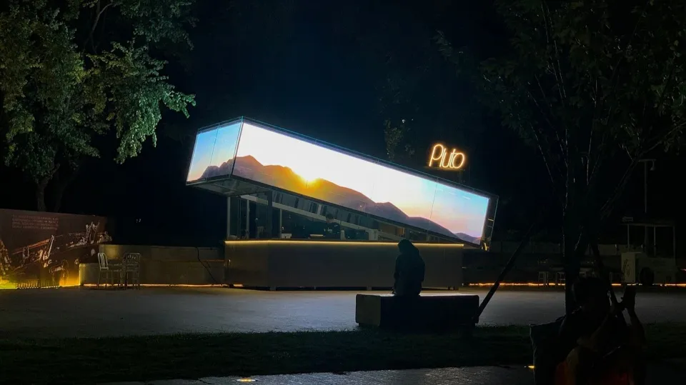 A nighttime building facade with a large window display showcasing a sunset view, accompanied by two benches and a calm atmosphere
