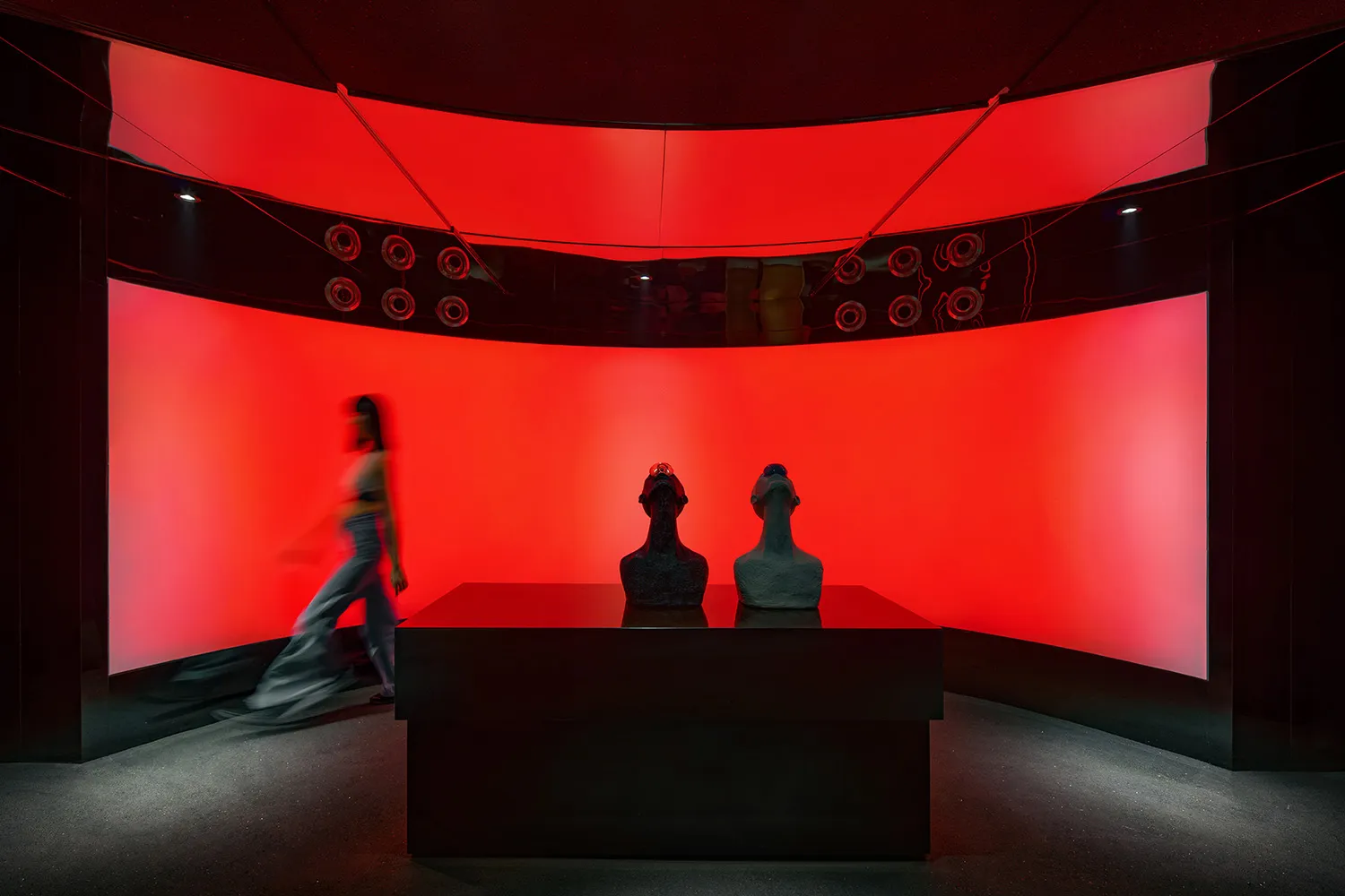 A modern gallery space with red walls, black tables, and a display of three sculptures on a central table