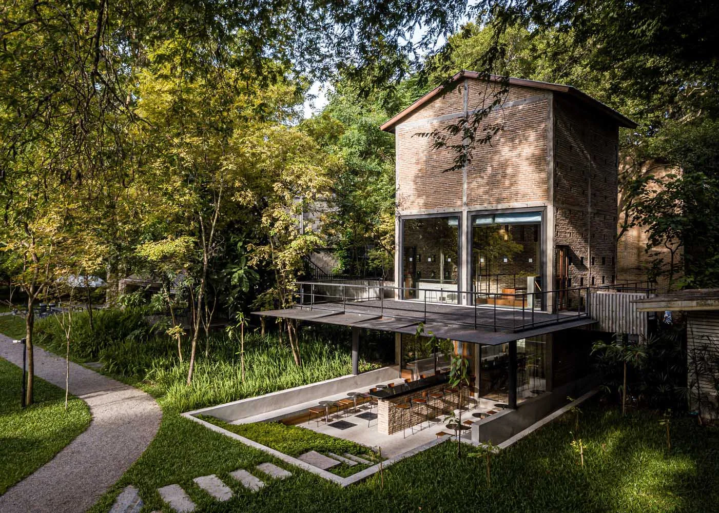 A house with a brick exterior and large windows surrounded by trees in a forest, featuring chairs and potted plants