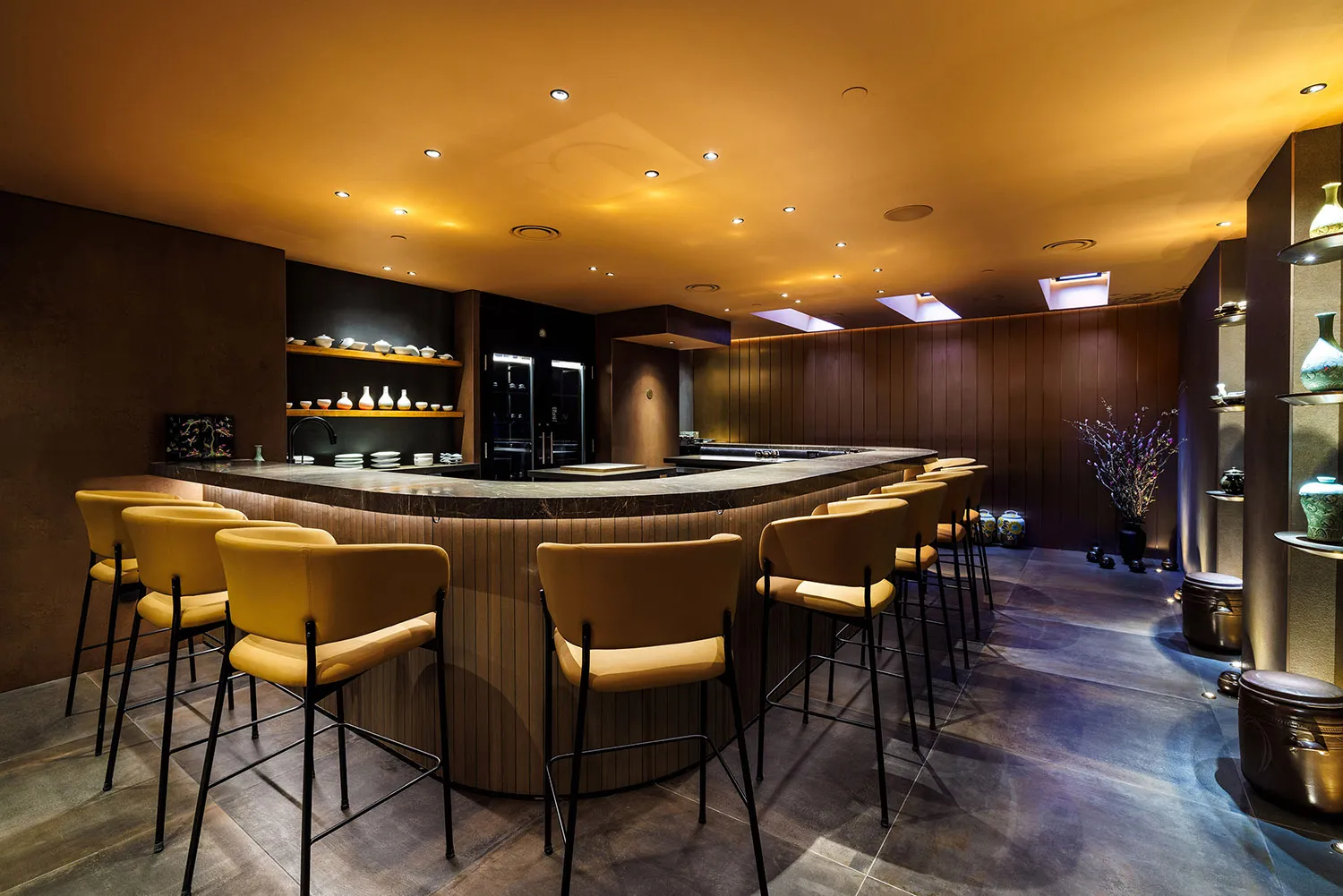 A modern restaurant with a large bar area featuring a wood countertop and metal stools, surrounded by chairs and tables in a warm and inviting atmosphere