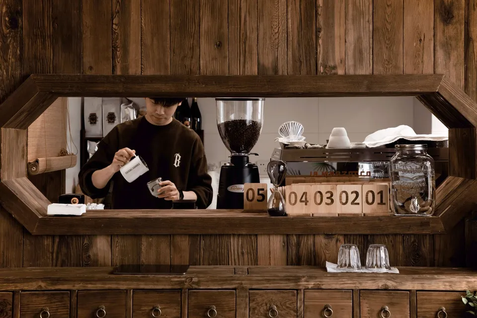 A man prepares coffee at a wooden counter with multiple cups and bottles nearby, in a casual and inviting atmosphere