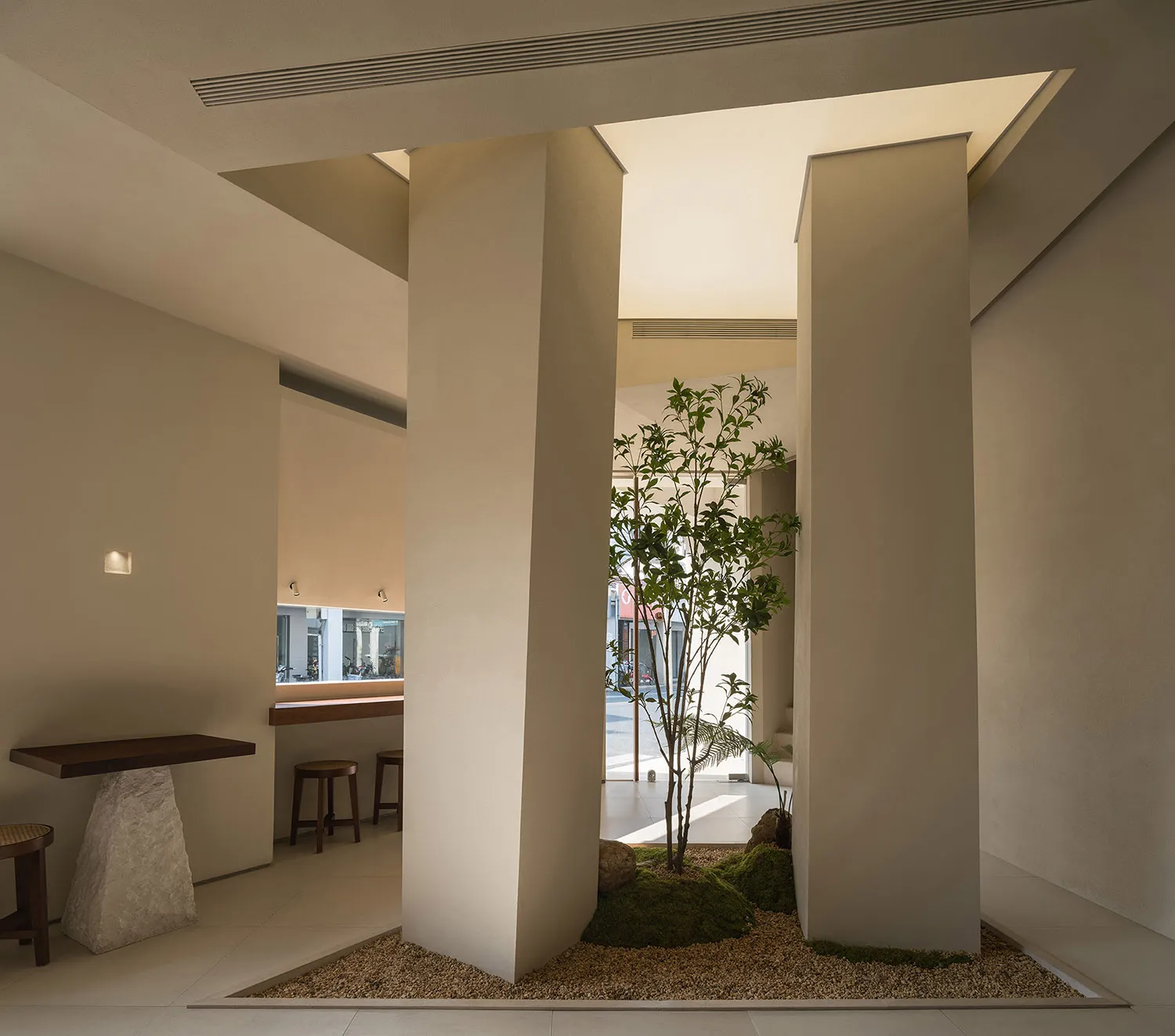 A calm and inviting indoor space with a tree growing through a large white wall, featuring two chairs and a dining table, surrounded by greenery