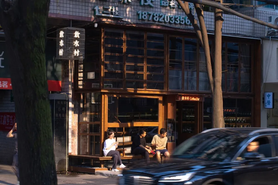 An Asian restaurant with people sitting outside on wooden benches in front of the establishment, surrounded by glass walls and natural light