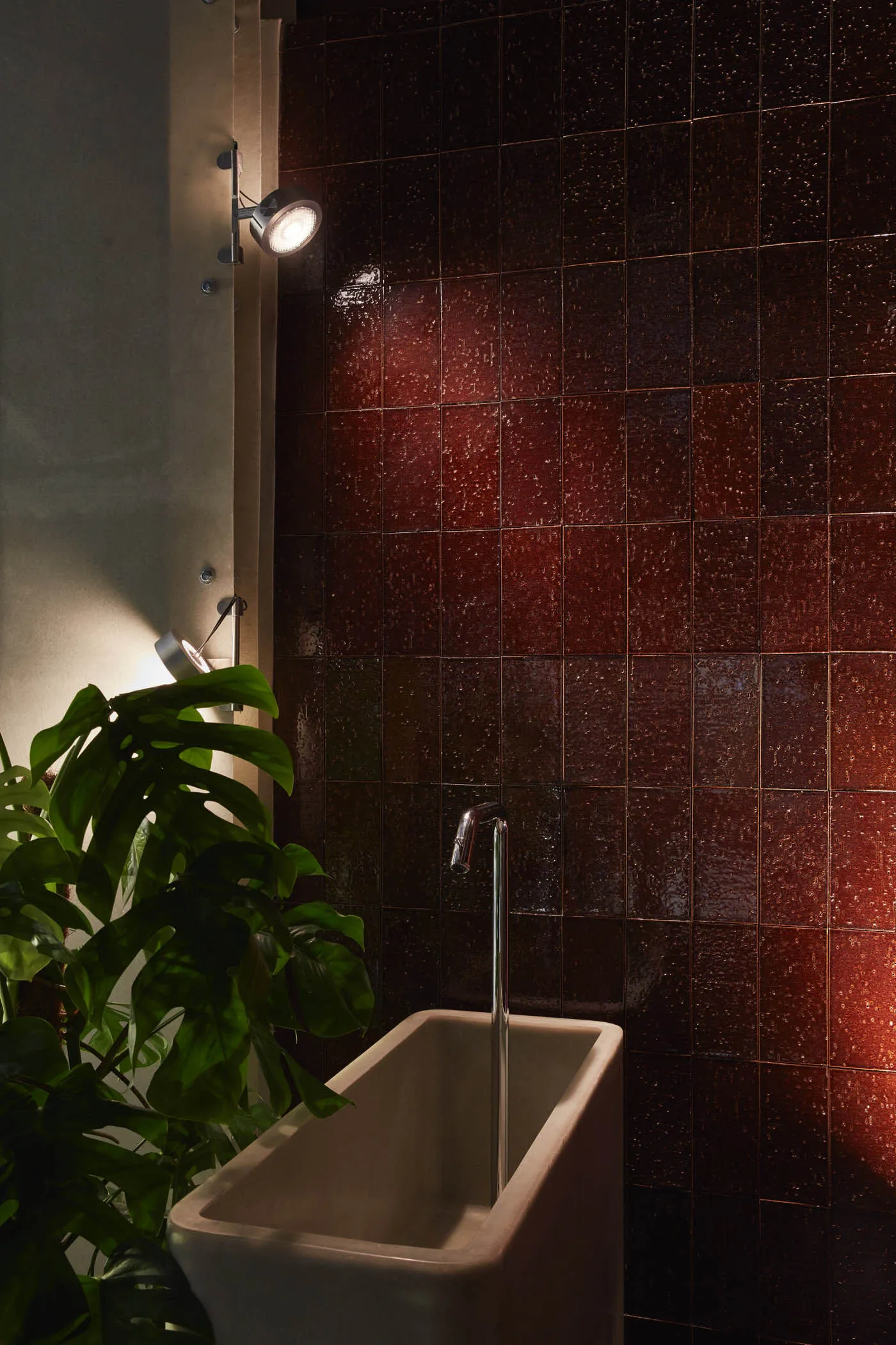 Bathroom with a white porcelain sink, red wall tiles, and a potted plant, illuminated by overhead lights