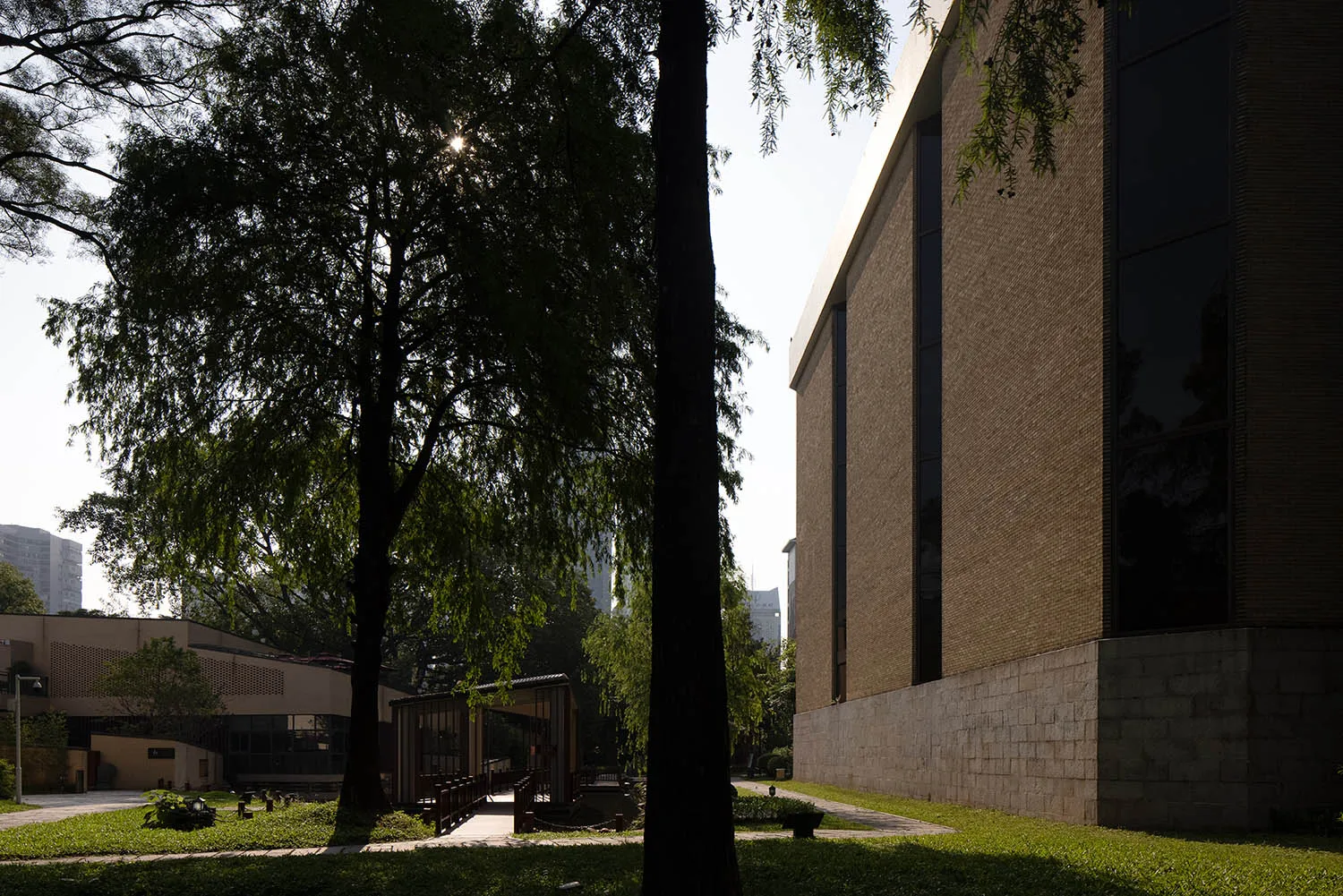 A modern building with a brick facade, surrounded by trees, with sunlight illuminating the scene, creating shadows and highlights on the building's surface