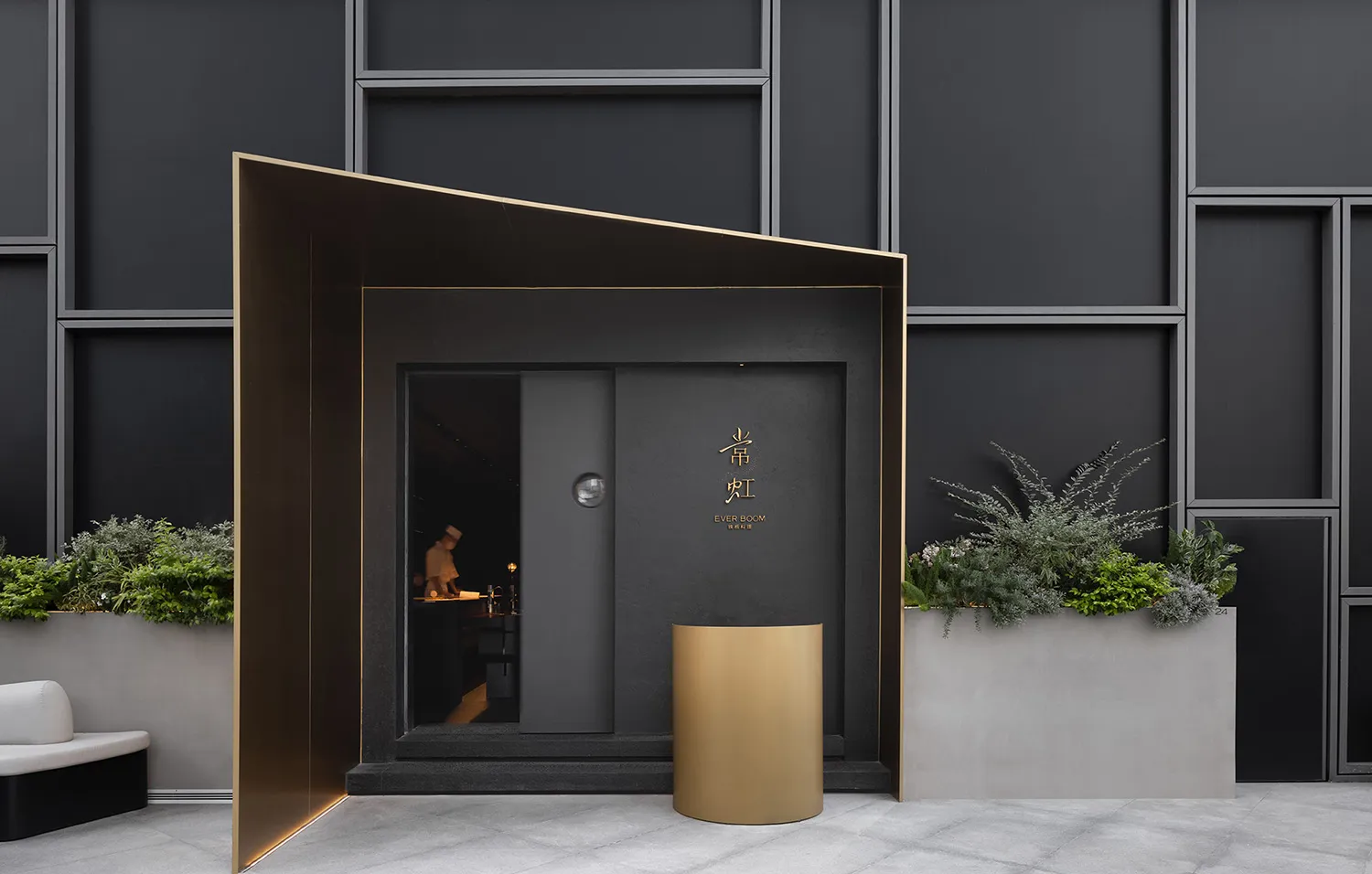 A restaurant entrance with a black door, gold trim, and a dining area with a table and chairs, surrounded by a potted plant