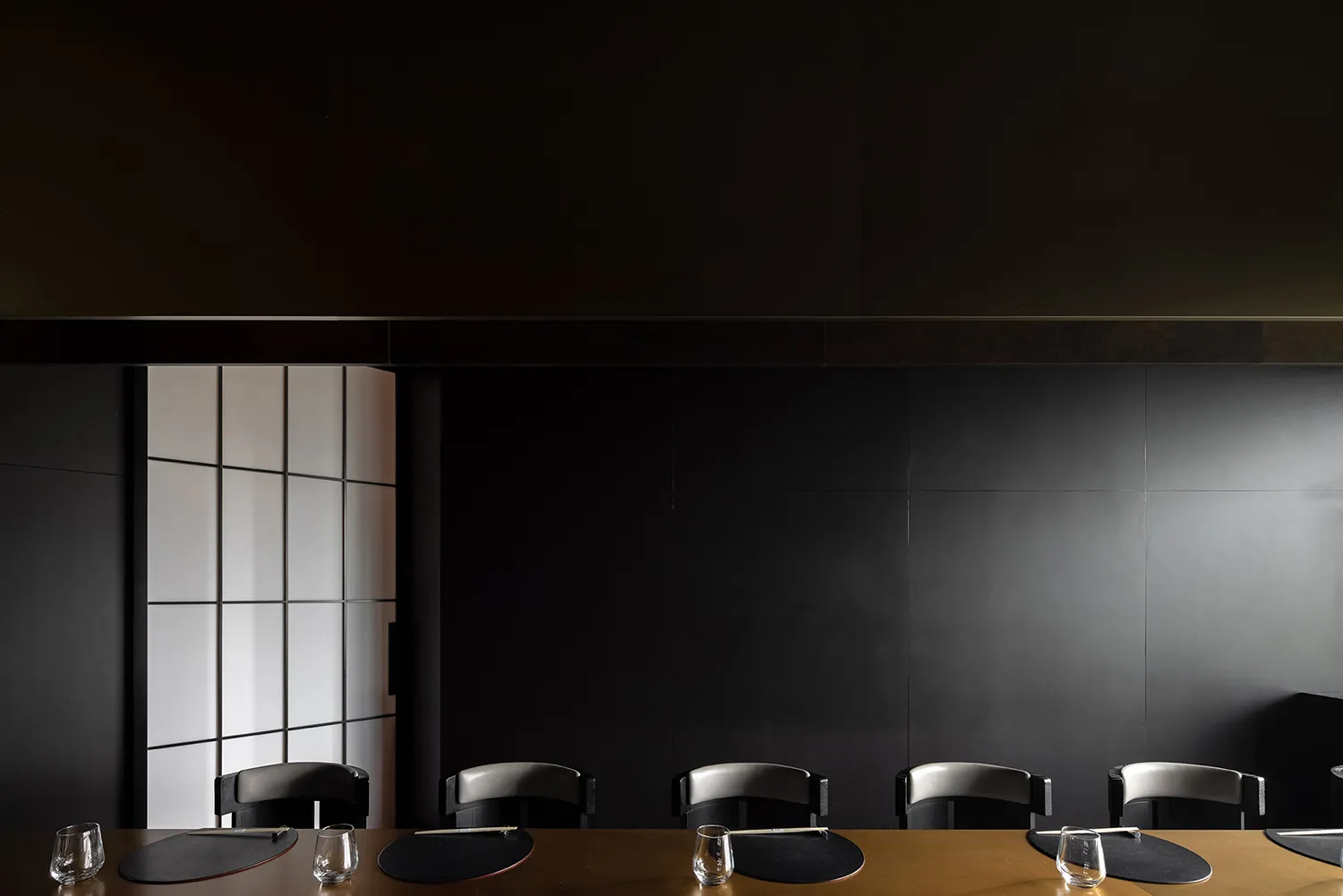 A modern dining area with a long black table and multiple chairs, set for a formal dinner with wine glasses, cups, and utensils