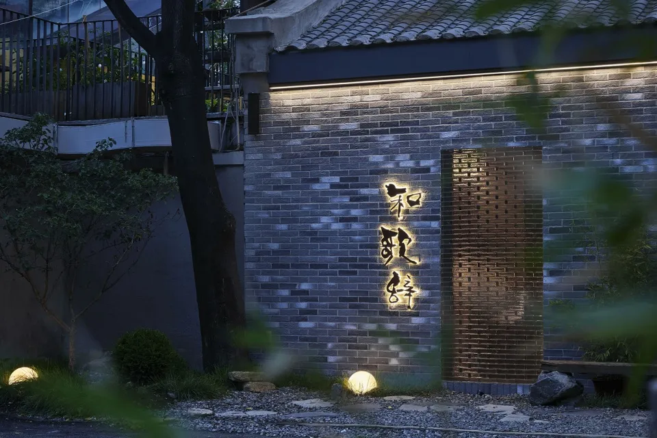 Asian-style building with a brick wall and stone facade, featuring a doorway with Chinese characters, surrounded by two trees in a calm and serene atmosphere