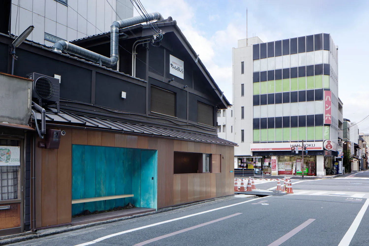 A modern building with a wooden facade and a blue roof, featuring a small shop or cafe in front, outdoor benches, and construction barriers