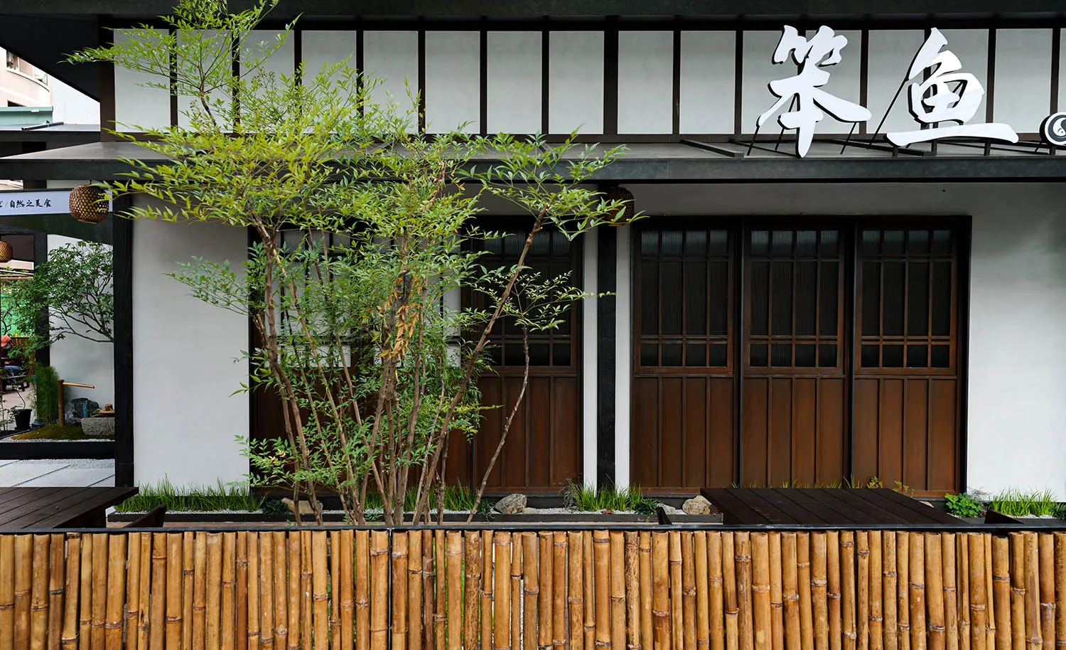 Asian-style building with wooden walls, bamboo fencing, a balcony, and a doorway leading to a patio area, surrounded by two potted plants