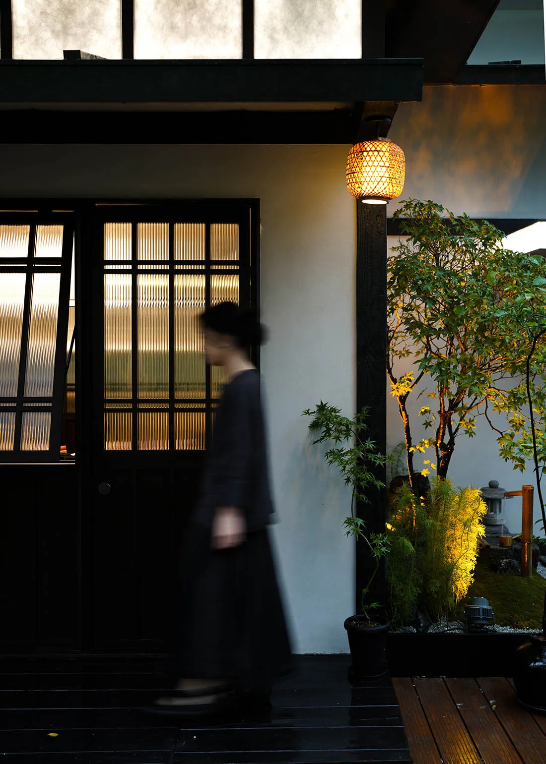 A woman in a black dress walks down a hallway with a white wall, a potted plant, two vases, and a bowl