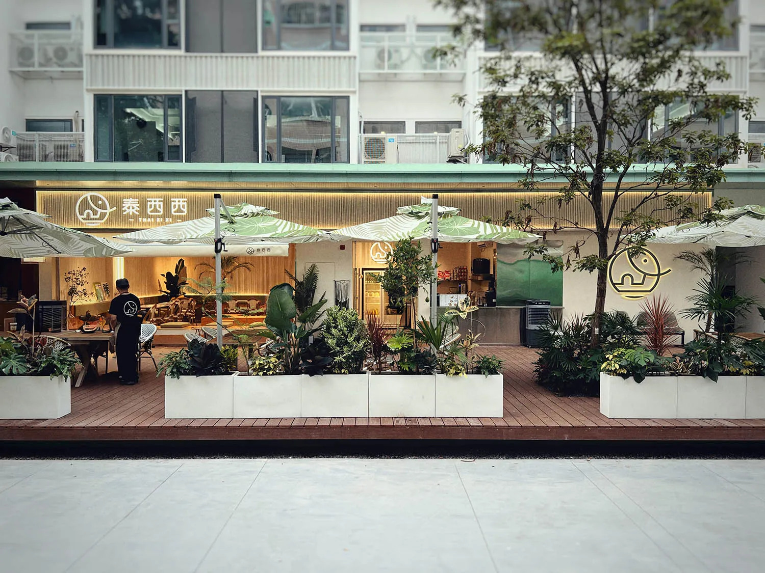 An outdoor cafe with umbrellas, potted plants, chairs, and dining tables, set against a background of refrigerators and a pleasant atmosphere