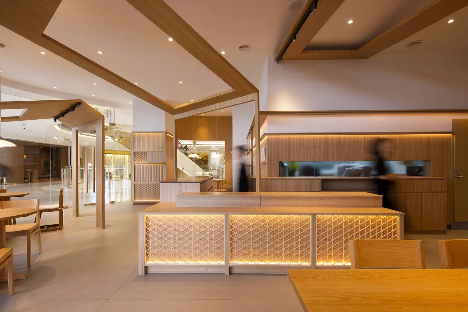 Open dining space with wood elements, two dining tables, and a woman walking through the room, with a bright and inviting atmosphere