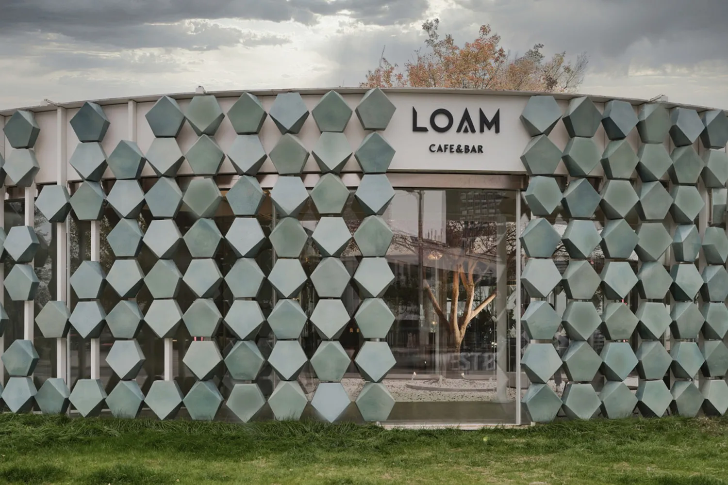 A modern cafe with a glass wall featuring hexagonal shapes, surrounded by a tree and outdoor seating area