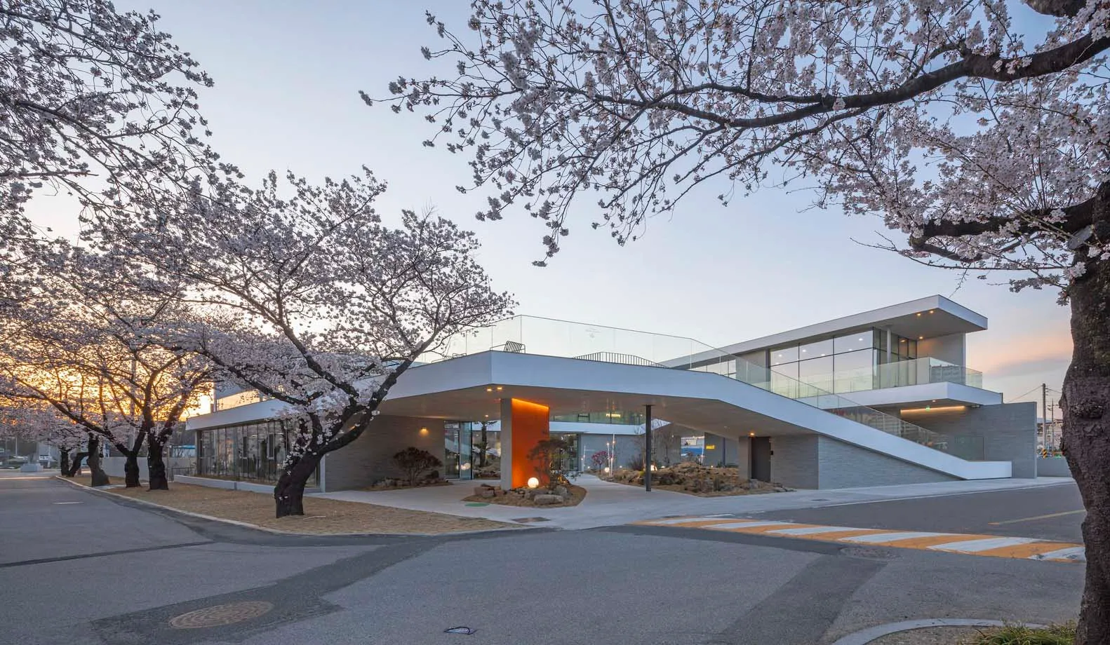 Modern building with a large glass wall, surrounded by trees and flowers, featuring a curved roof and a serene atmosphere