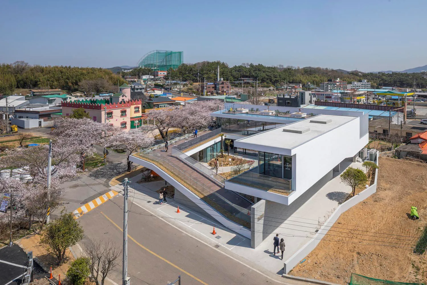 A modern building with a white exterior and a large glass wall, featuring a bridge connecting two parts and people walking around the area