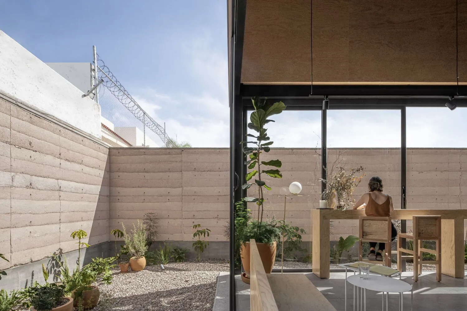 A well-lit living area with a large window, dining table, chairs, potted plants, and a bench, featuring a mix of natural and artificial light sources and a modern, contemporary aesthetic