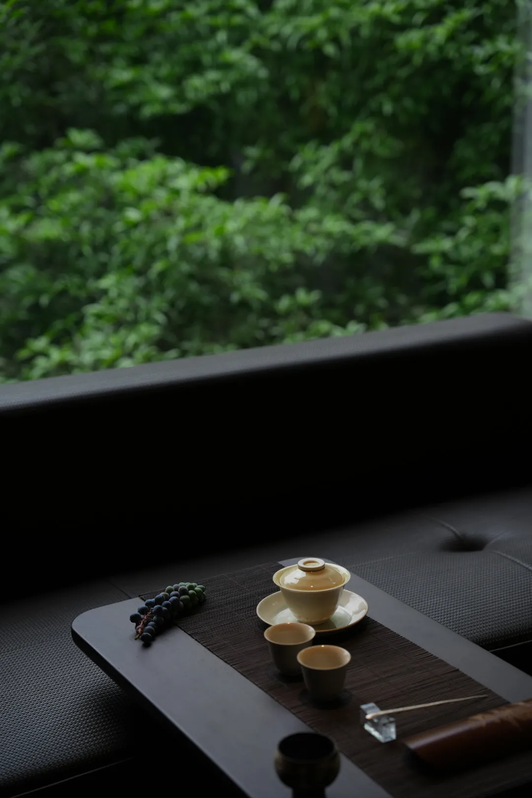 Cozy living space with a black sofa, natural light from a window, and a table with cups, a bowl, and grapes