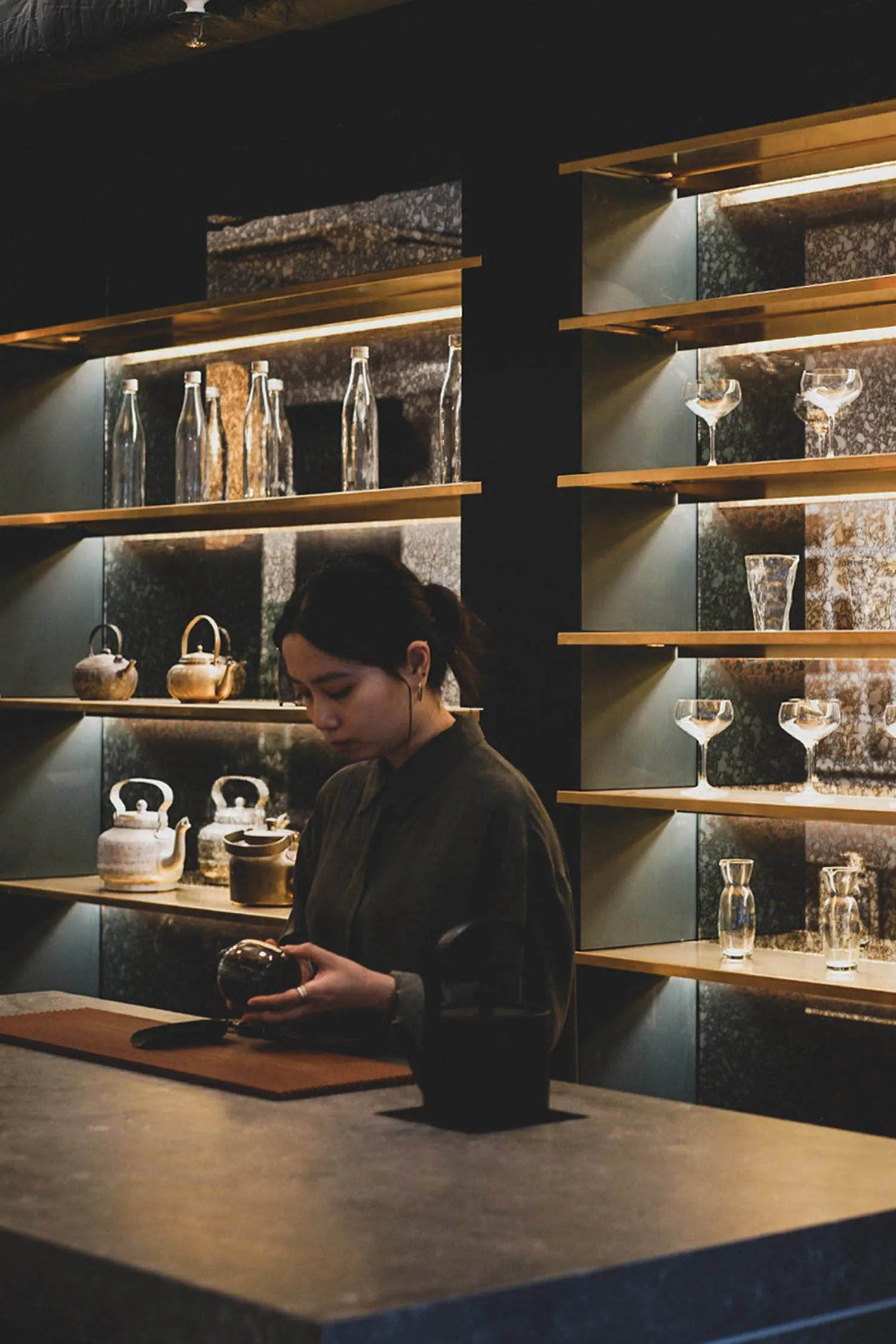 A woman stands in front of a wooden counter filled with wine glasses, cups, and bottles, surrounded by a social gathering atmosphere