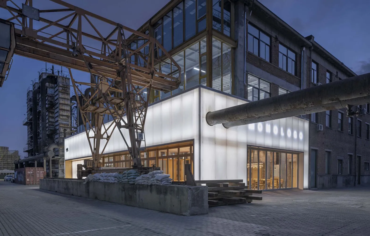 A modern building with a white glass facade and windows, featuring a large crane and chairs in the foreground, set against a night-time urban backdrop