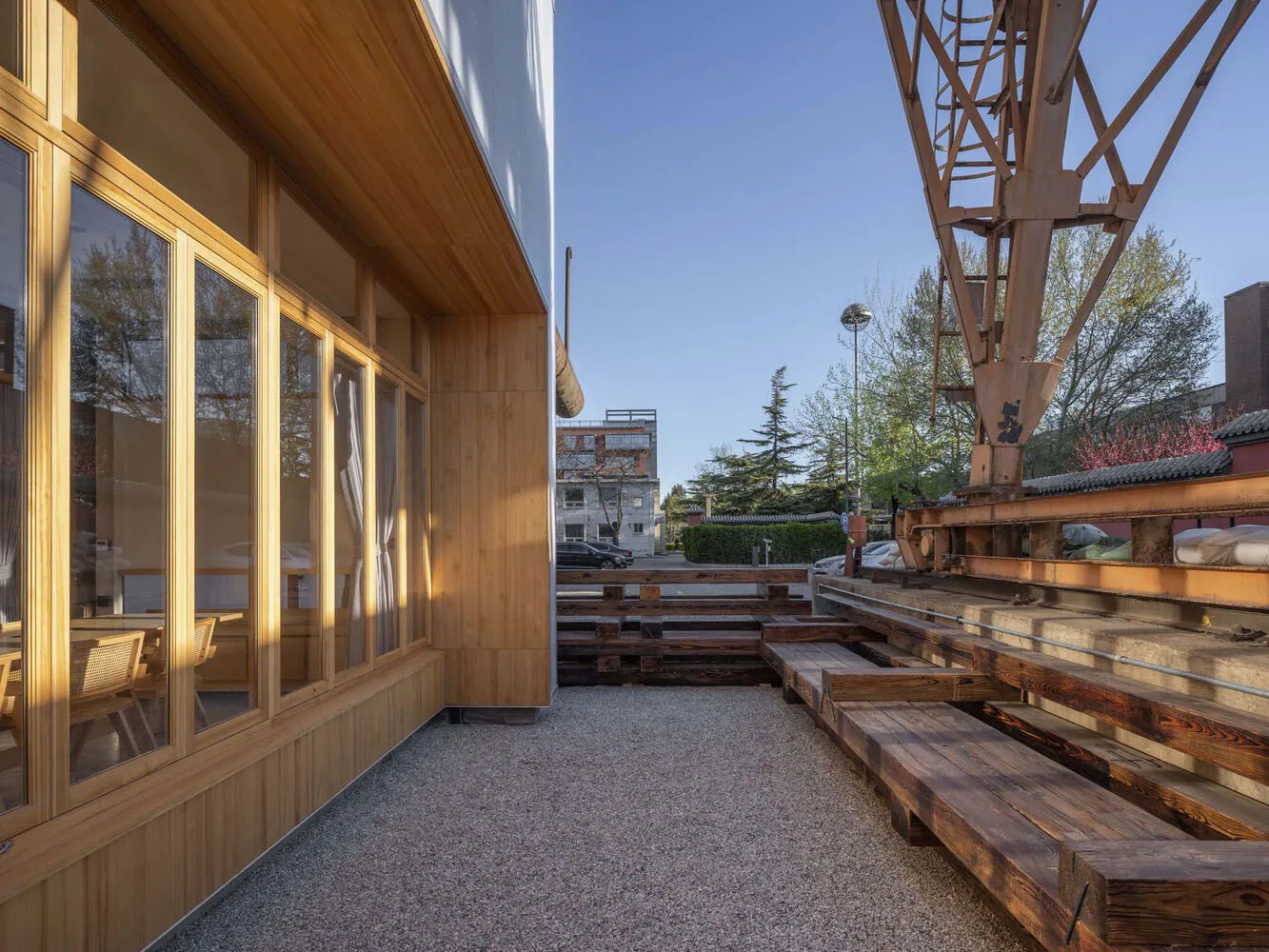 A modern building with wooden walls and a cement ground, featuring large windows and a contemporary design, set against an urban backdrop with parked cars