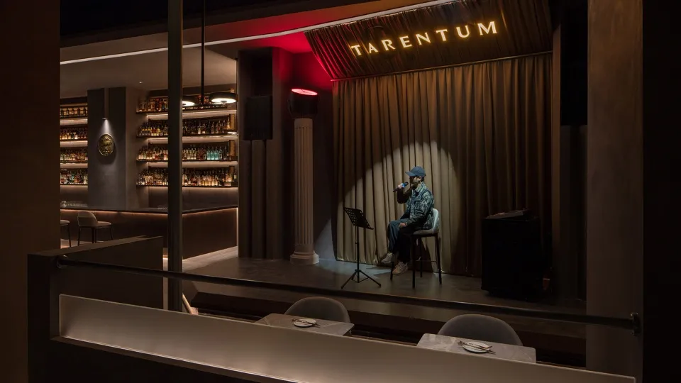 A man sings into a microphone on a stage, surrounded by chairs and bottles in a dimly lit room
