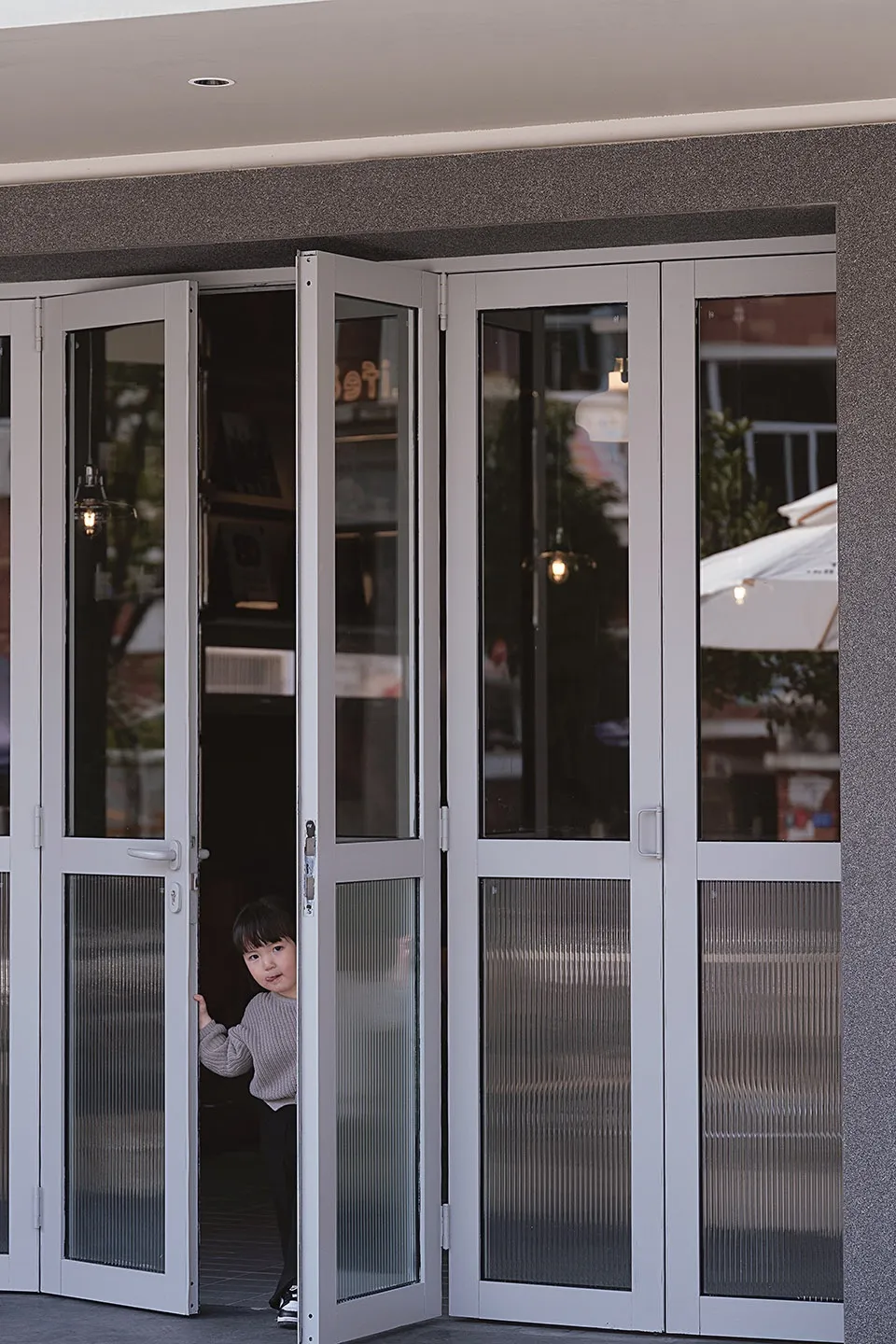 A young child stands in front of a glass door with a silver frame, peeking through the sliding doors with a curious expression
