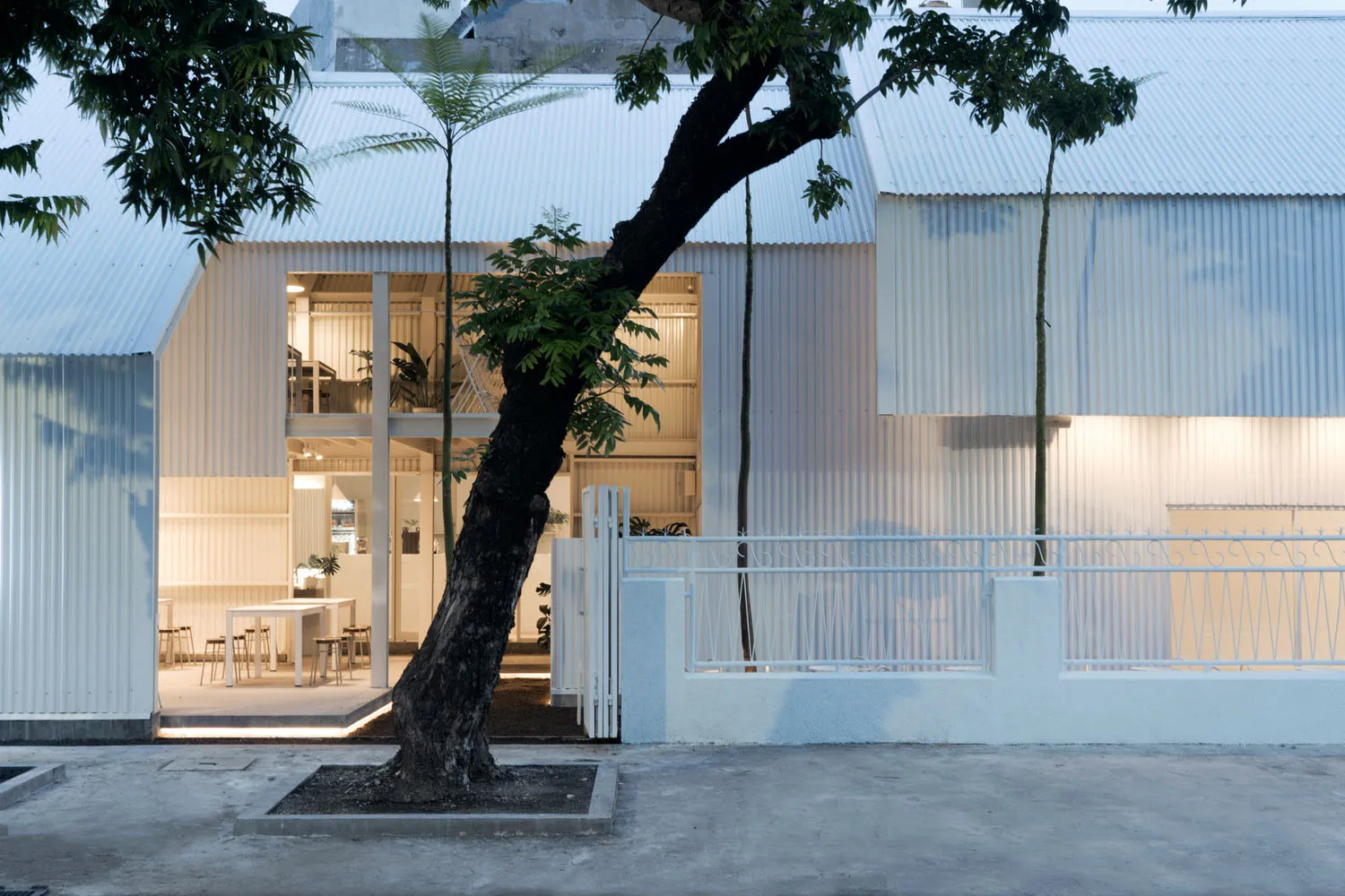 A modern outdoor space with a white metal building and a large tree in front, featuring several chairs and two dining tables