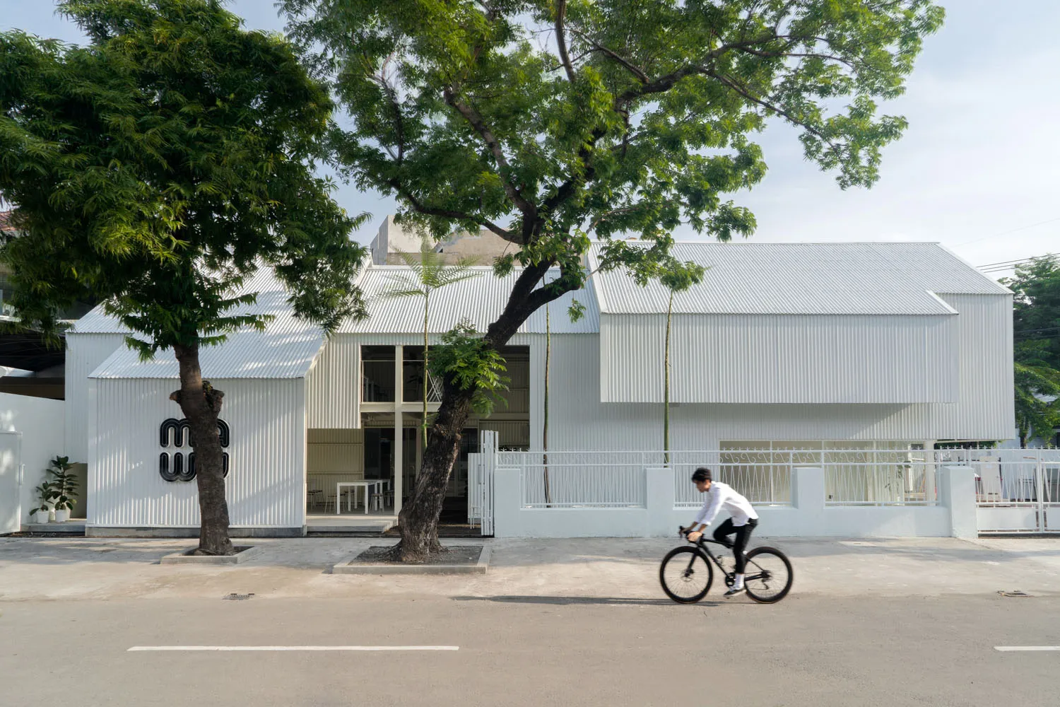 A modern building with a white concrete facade, featuring outdoor seating areas with two chairs and a bench, set in a calm and peaceful environment