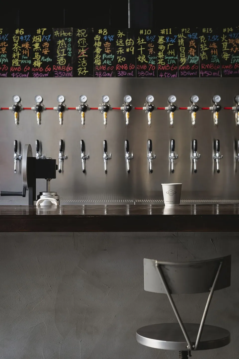 Industrial-style bar area with a long counter featuring 14 beer taps and two cups, set in a busy restaurant or bar atmosphere