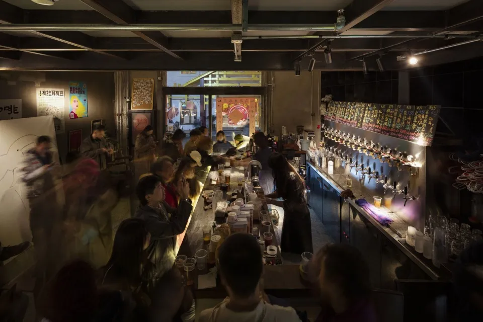 A lively bar scene with people gathered around a counter, enjoying drinks and socializing, surrounded by bottles, cups, and bowls, with a casual and relaxed atmosphere