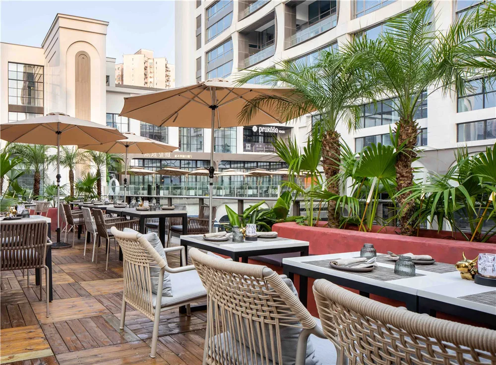 A dimly lit dining area with multiple tables and chairs, concrete walls, and umbrellas providing shade