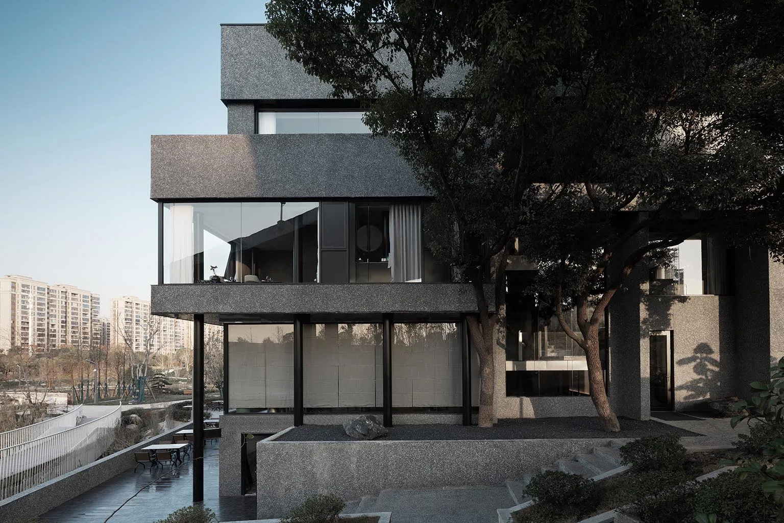 A modern building with gray stone walls and black glass windows, set against a backdrop of trees in an urban environment