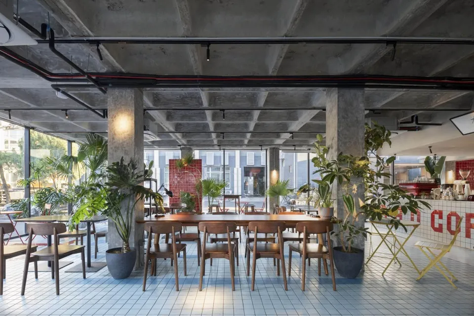Modern restaurant interior with a large dining table, red accent, and potted plants, surrounded by chairs in a warm and inviting atmosphere