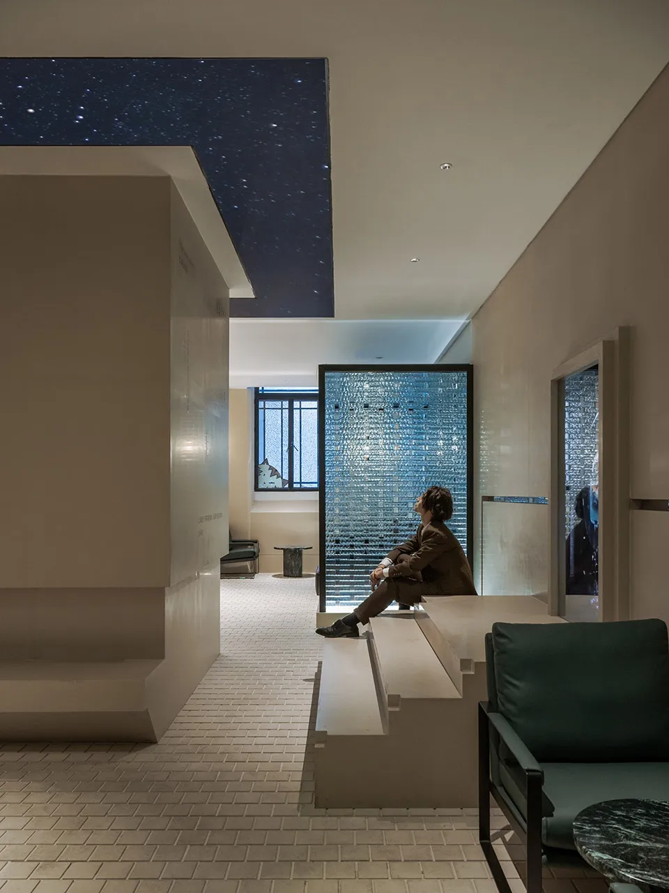 A modern living area with a man sitting on a bench in front of a window, surrounded by two chairs and concrete walls that allow natural light to enter