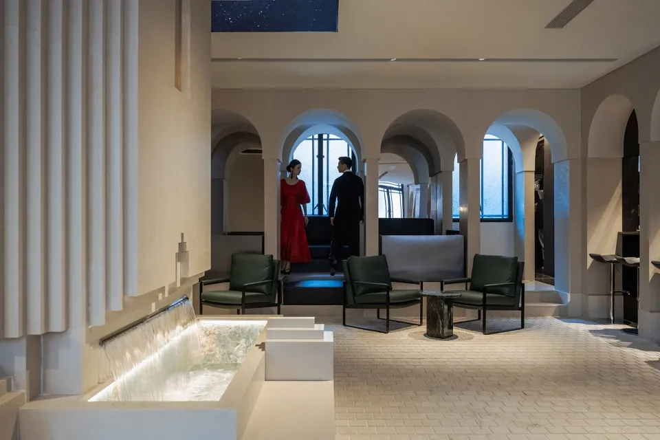 Spacious and luxurious interior design with high ceilings, well-lit and featuring chairs, a bench, and two people standing in the room