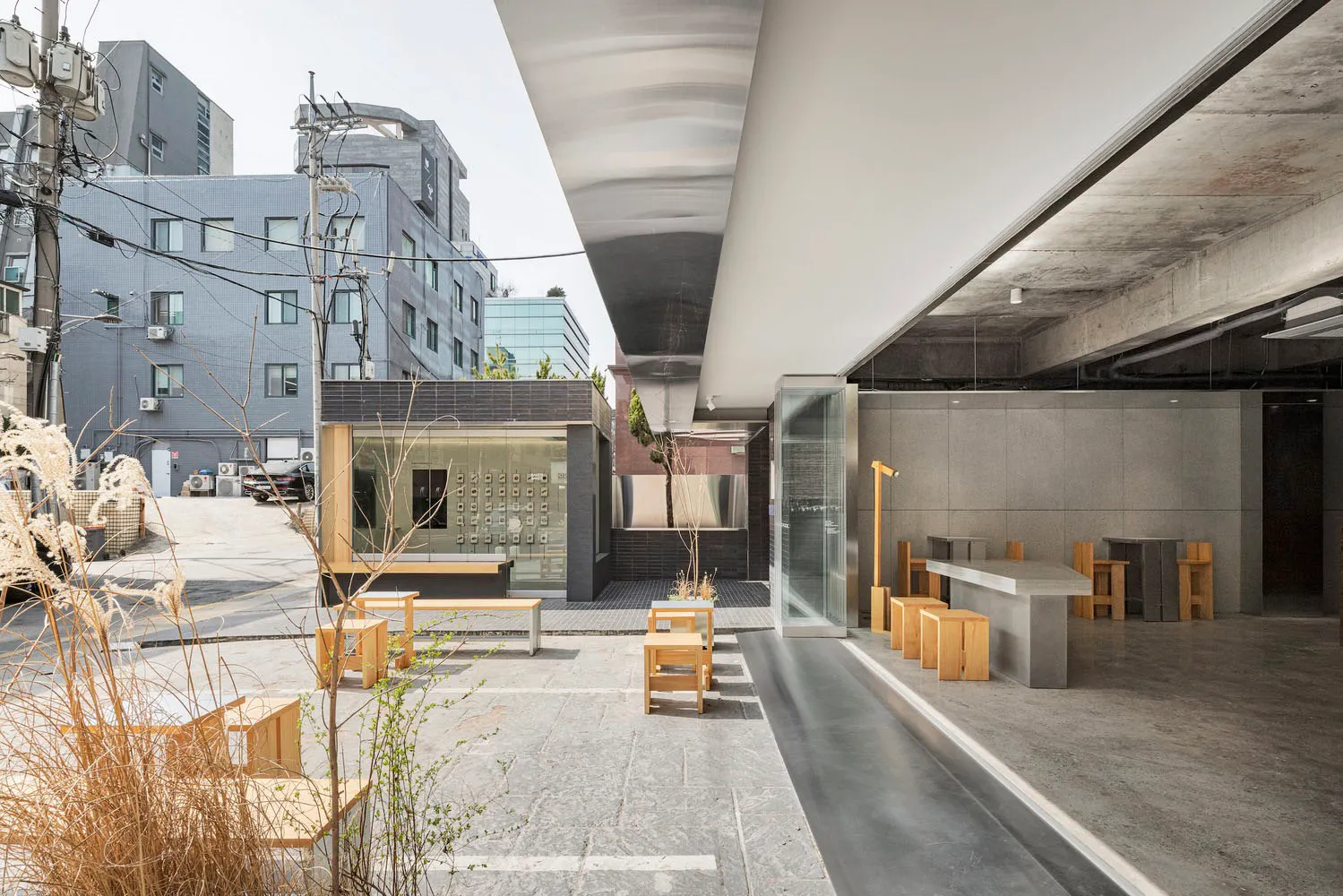 A modern building with a glass wall facade, wood and concrete elements, outdoor seating, and potted plants