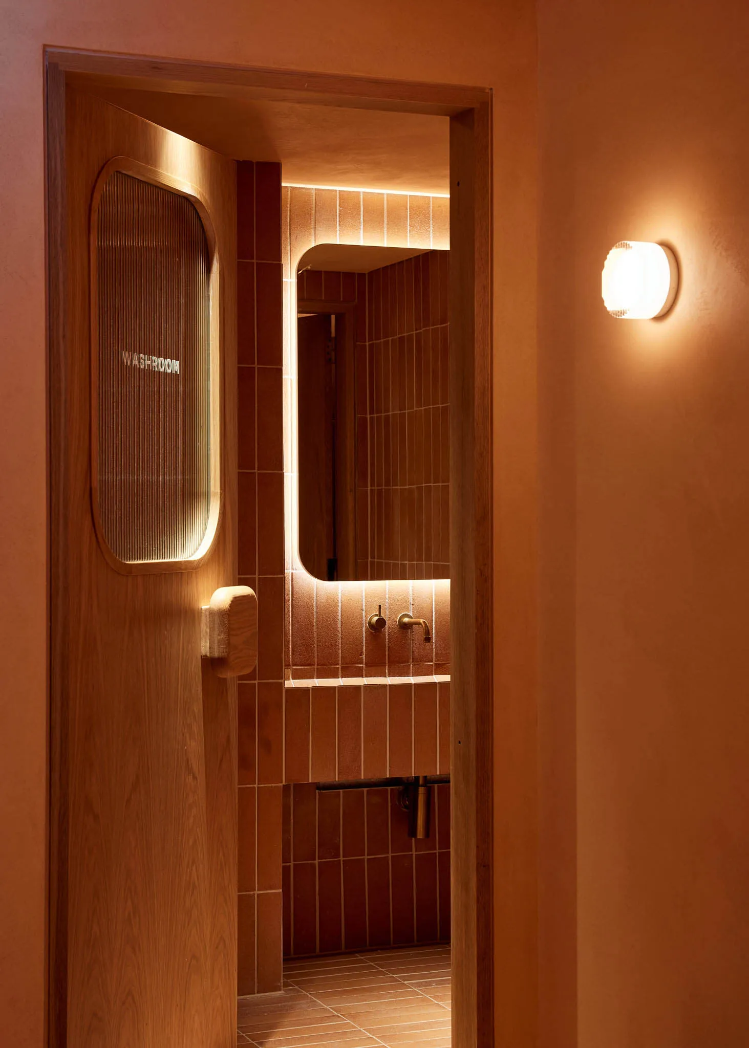 A simple bathroom with brown tiled walls, a sink, toilet, and mirror, creating a warm atmosphere
