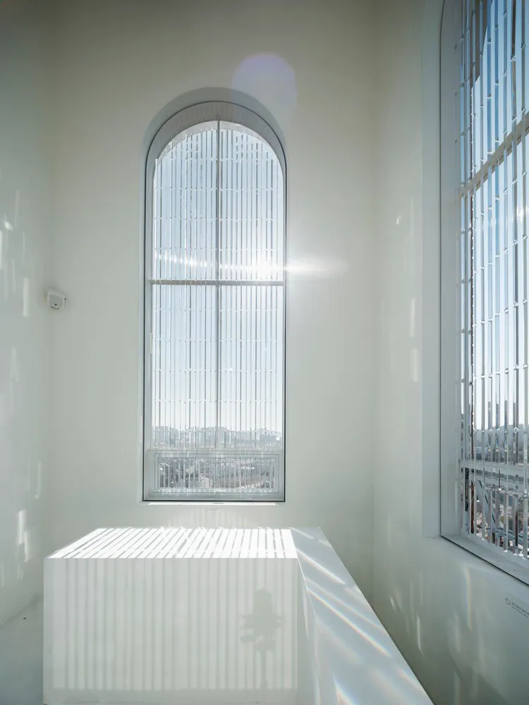 An empty white room with two beds and a chair, lit by natural light from a large window