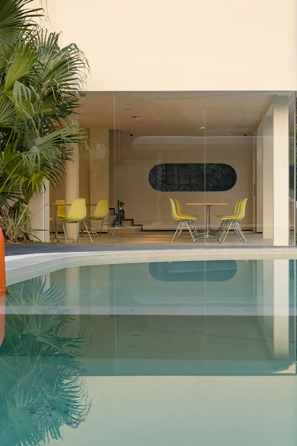 Indoor swimming pool area with a glass wall leading to a patio, surrounded by yellow chairs and a modern atmosphere