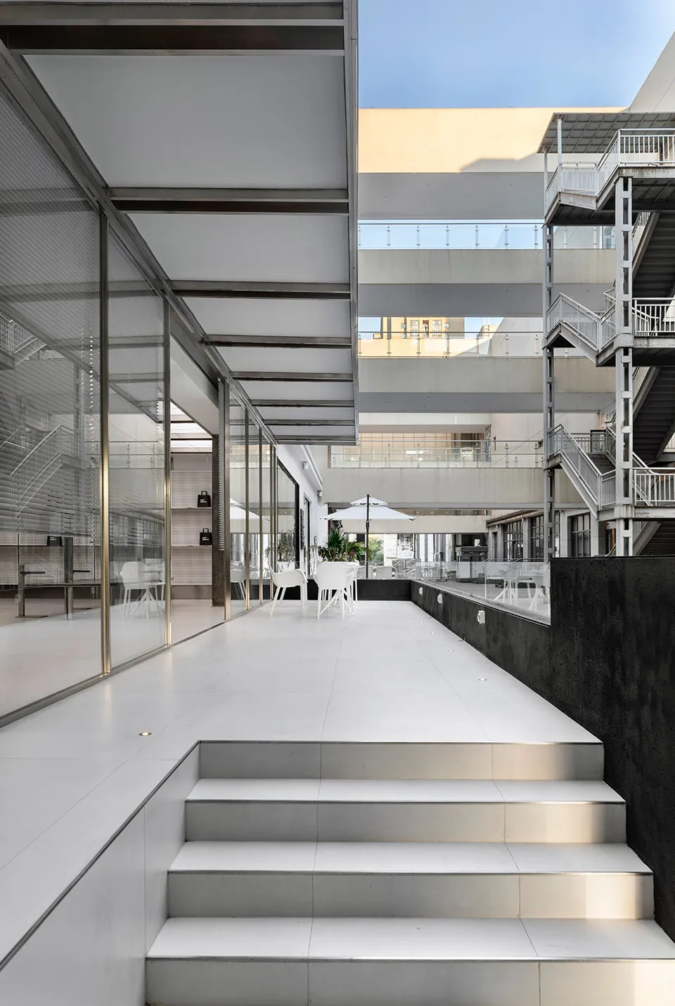 A modern indoor space with multiple levels, white walls, and a mix of chairs, stairs, and an umbrella, creating a well-lit and comfortable atmosphere