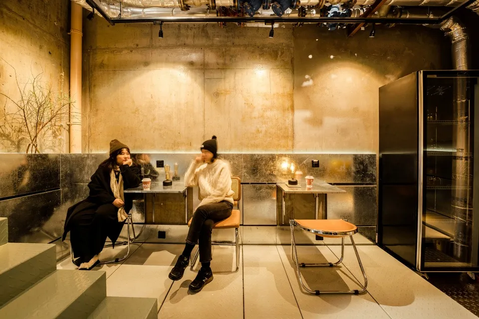 Two women sit in a dimly lit art gallery or indoor space with chairs, cups, and books, creating a cozy and intimate atmosphere